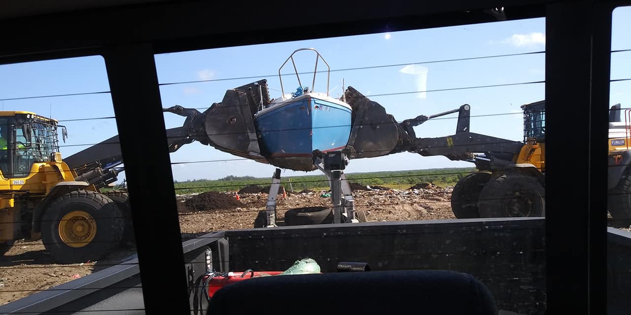 Heavy equipment lifting a blue sailboat hull at a disposal site