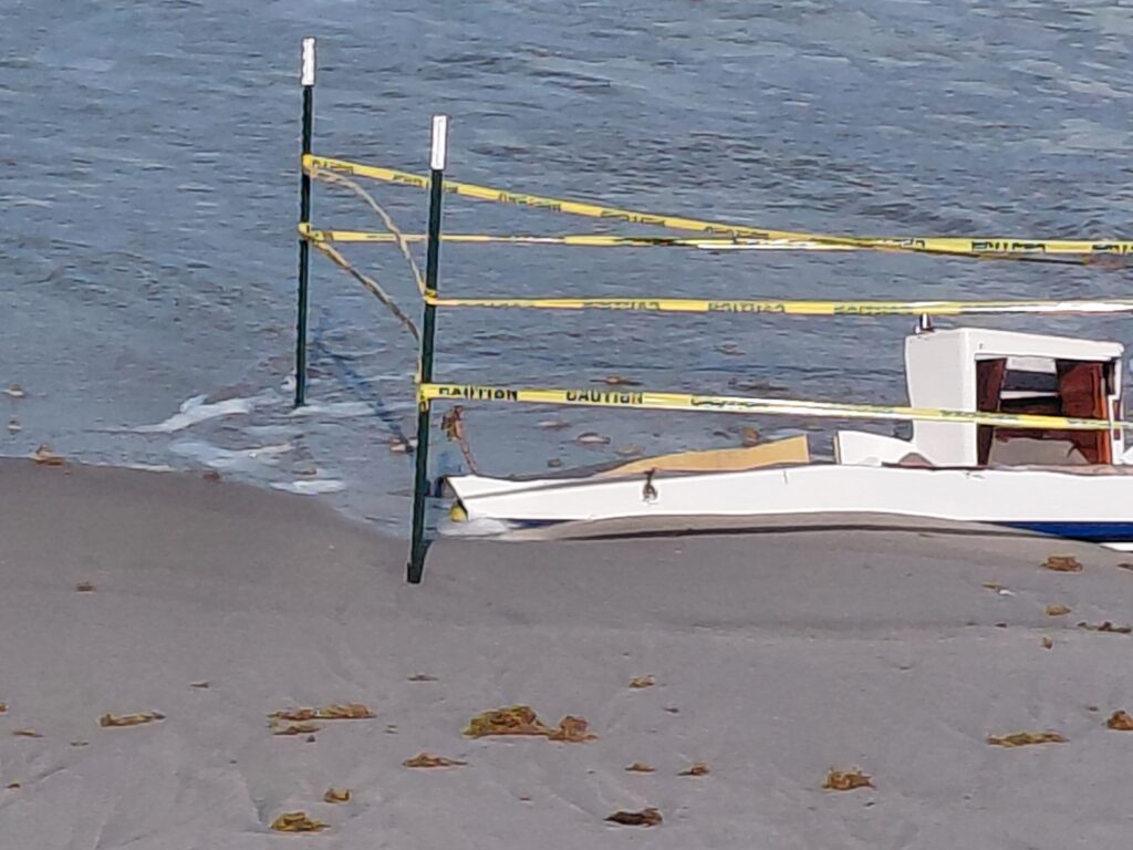 Separated boat sections behind caution tape along a sandy shoreline