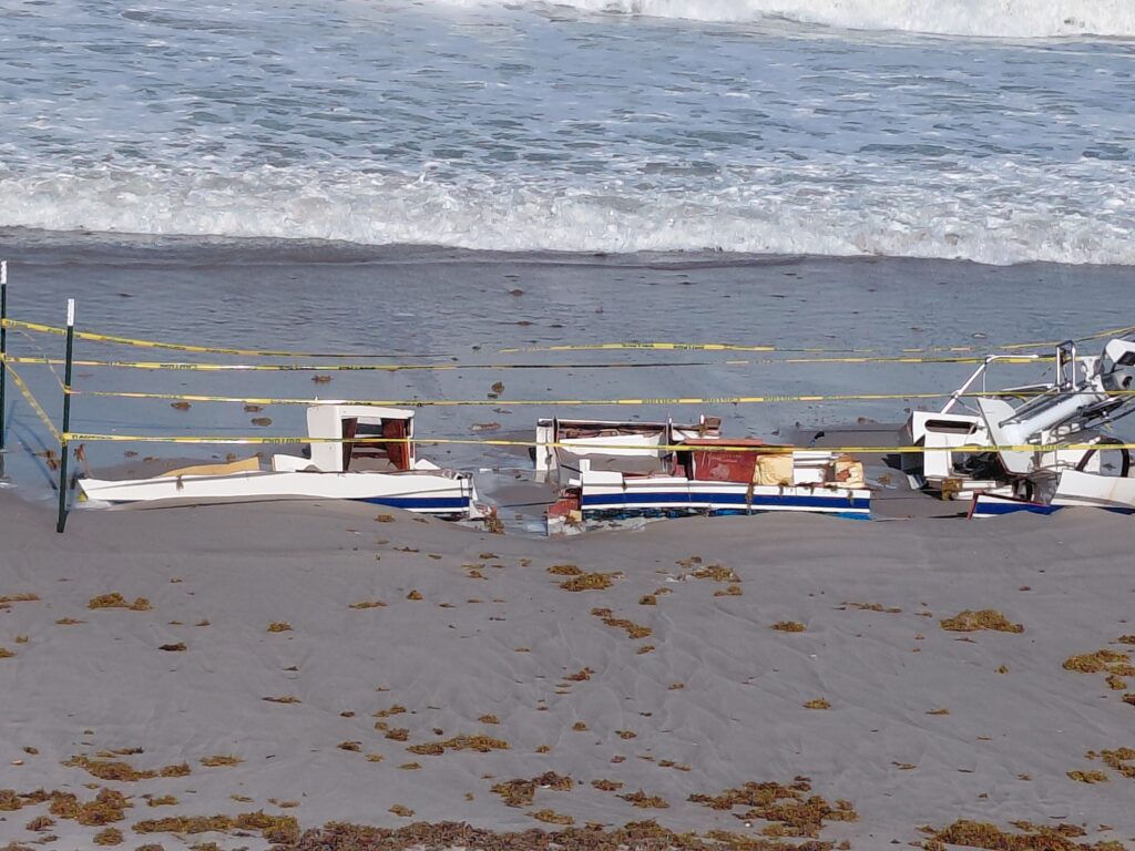 Broken vessel sections behind caution tape beside the surf