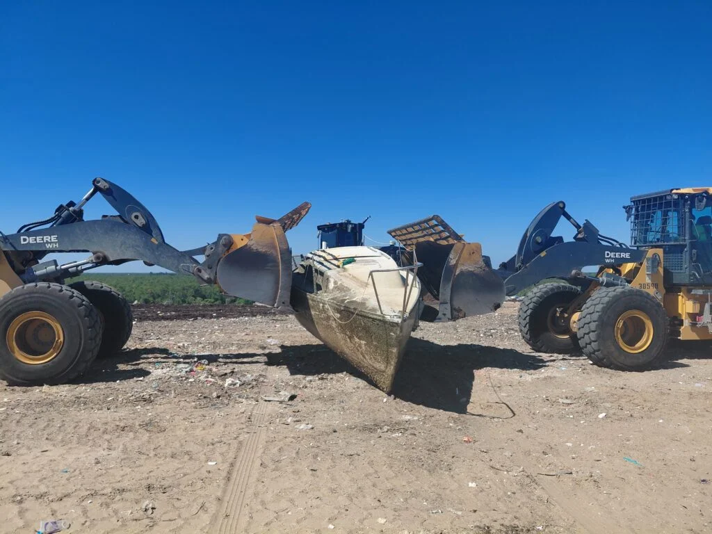 Two wheel loaders positioned beside a deteriorated sailboat hull