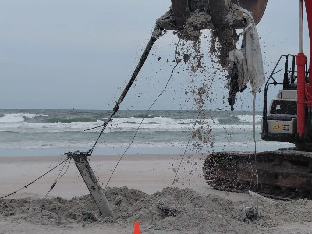 Excavator lifting vessel material from sand beside the ocean
