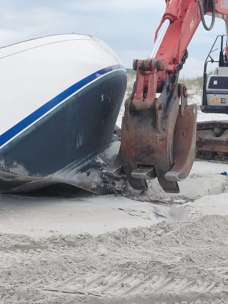 Excavator bucket beside a damaged boat hull resting on sand