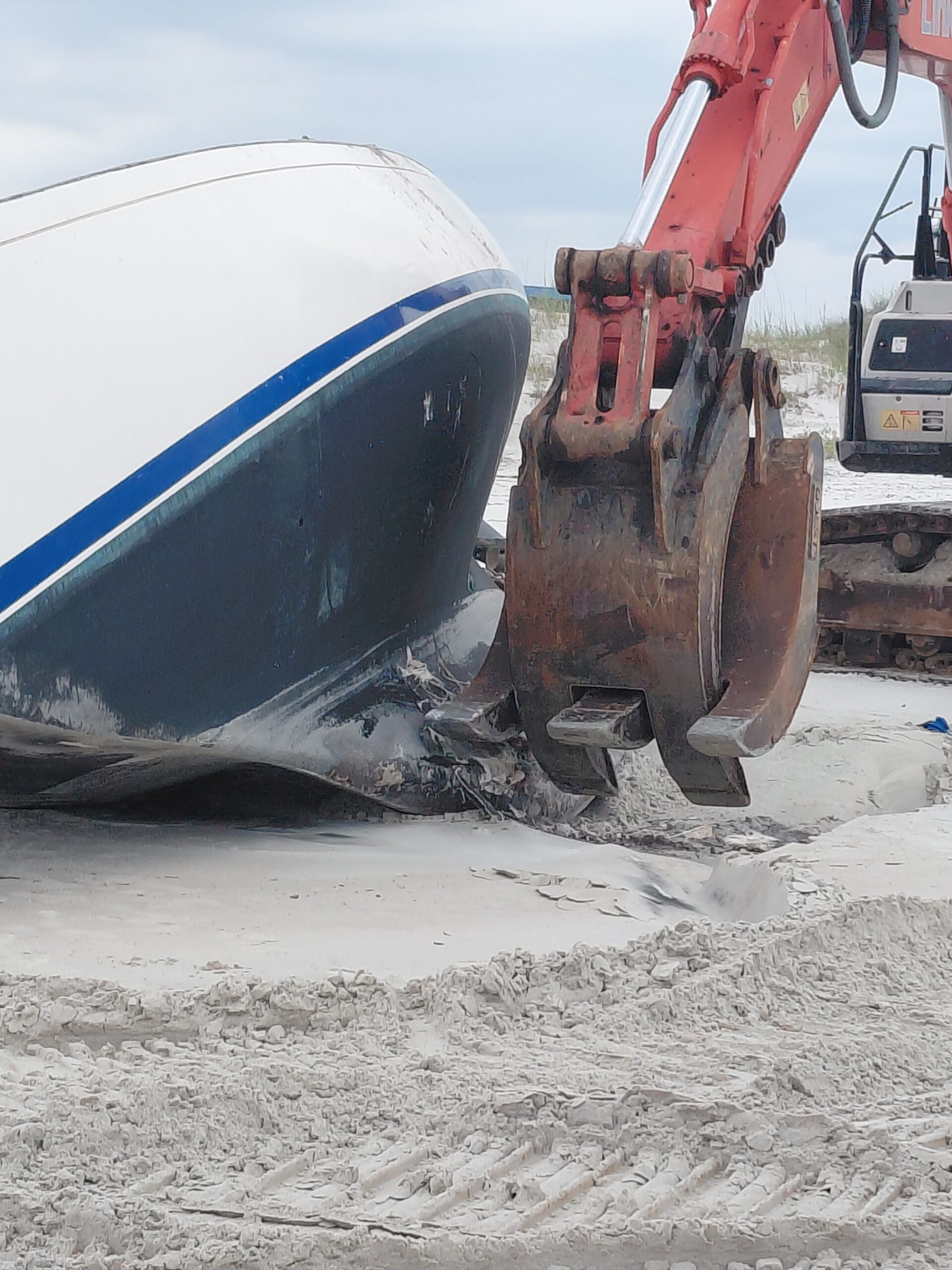 Excavator bucket beside a damaged boat hull resting on sand