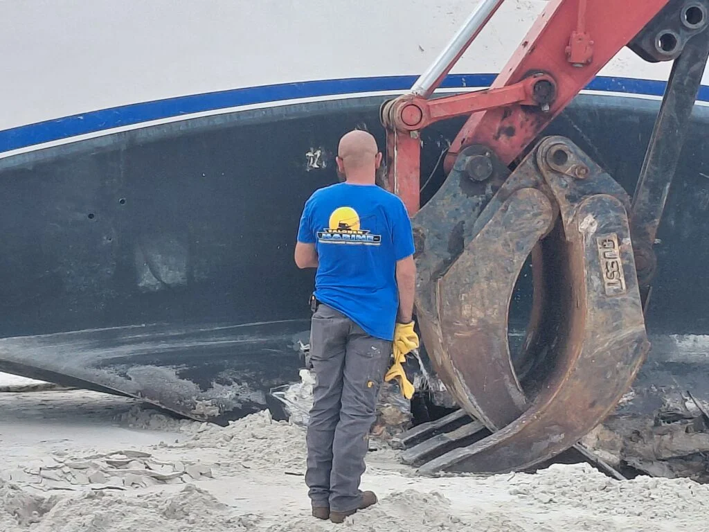 Worker beside a large boat hull and excavator attachment on sand