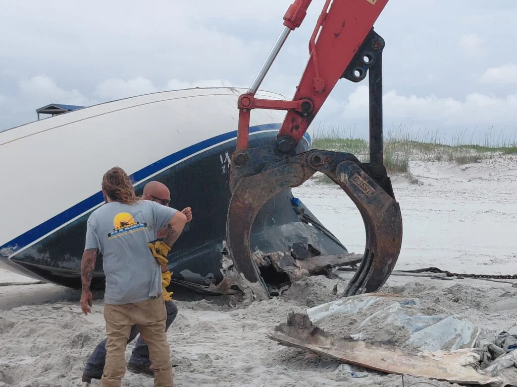 Crew and excavator beside a damaged boat hull on sand