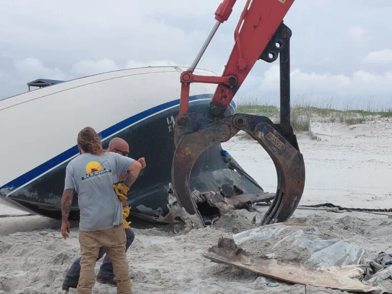 Crew and excavator beside a boat hull on a sandy shoreline