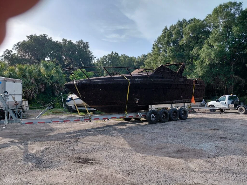 Damaged dark boat hull secured to a triple-axle trailer in an outdoor yard